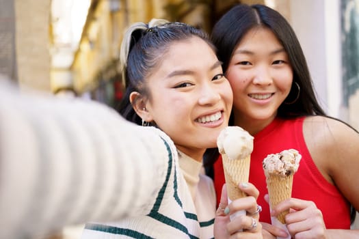 Positive Asian female friends taking self portrait while eating delicious ice cream cone against blurred background