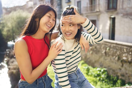 Happy young Asian woman sitting with hand on shoulder of cheerful girlfriend while spending time together in Granada