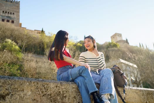 Happy young Asian female friends, in casual outfits smiling and looking at each other while talking on stone bench against ancient ruins of Paseo de los Tristes in Granada