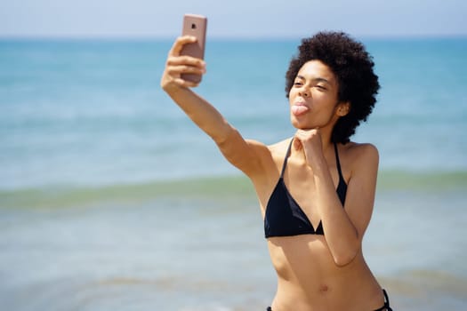 Funny African American female traveler in swimwear showing tongue and taking self portrait on smartphone while standing on coast near sea on sunny day