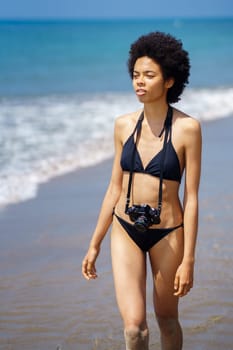 Serious African American female photographer with Afro hairstyle in swimwear with photo camera walking on shore near sea on summer day