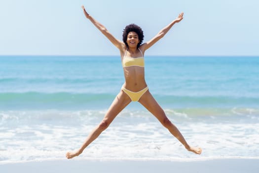 Full body of cheerful young fit African American female tourist, with dark curly hair in yellow bikini doing star jump, and smiling on sandy beach near wavy sea
