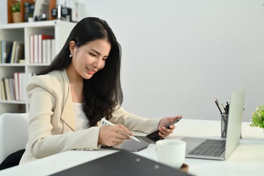 Attractive asian woman small business entrepreneur holding smart phone and using laptop at modern workplace.