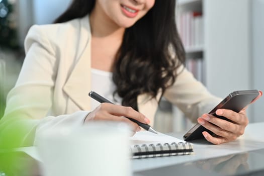 Smiling young businesswoman holding phone making important notes, planning daily appointment and write business trip in diary.