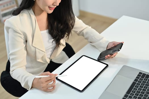 Cropped shot view of young businesswoman hand holding mobile phone and using digital tablet at office desk.