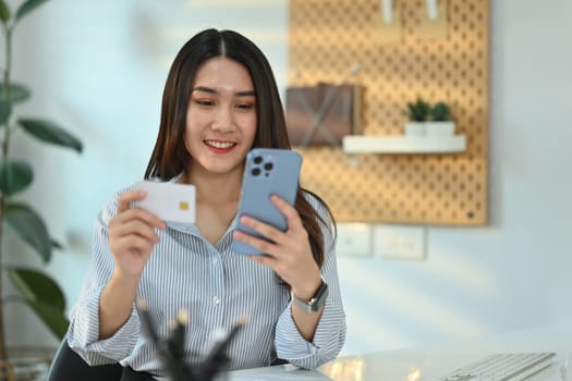 Smiling young woman holding credit card and smartphone, paying online, entering information, shopping online.