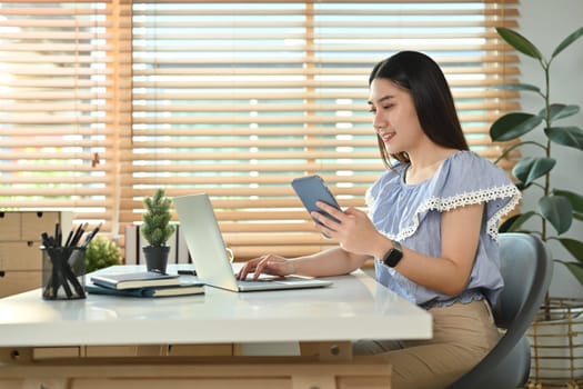 Attractive asian woman freelancer holding smart phone and using laptop at modern home office.