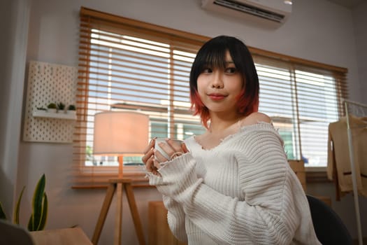 Relaxed young asian woman in knitted sweater holding cup of coffee and looking aways, sitting in cozy home.