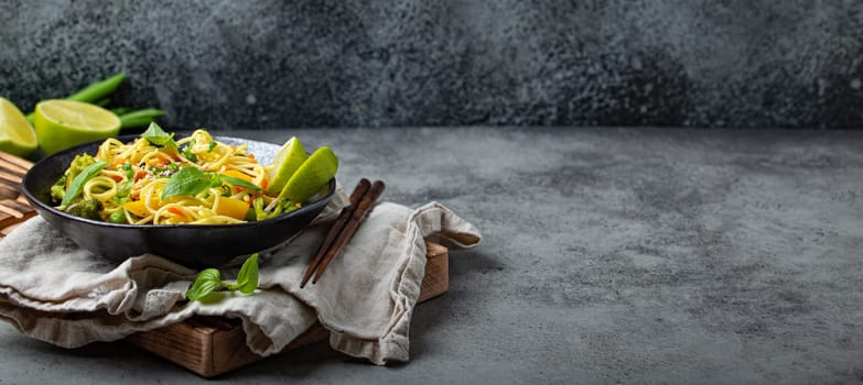 Asian vegetarian noodles with vegetables and lime in black rustic ceramic bowl, wooden chopsticks on cutting board angle view on stone background. Cooking noodles concept, copy space