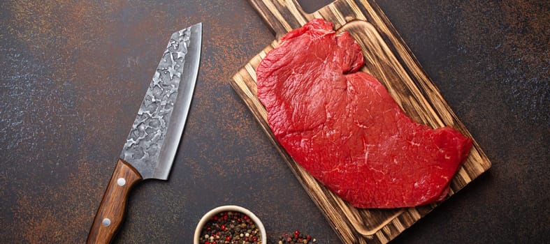 Raw uncooked top round beef steak on wooden cutting board with big kitchen knife and pepper on dark brown rustic stone background top view, cooking meat steak concept