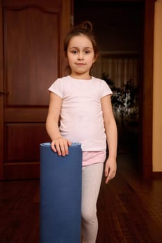Caucasian lovely 5-6 years child girl in sportrwear, holding gym mat and looking confidently at camera Sport. Yoga practice. Fitness concept