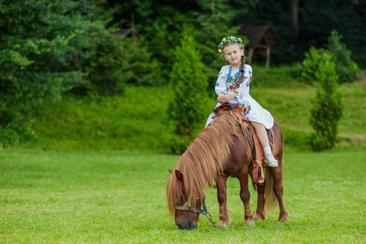 A little girl in the Ukrainian national costume rides a pony on the lawn