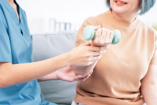 Caregiver helping contented senior woman exercise with dumbbell at home. Rehab for elder, senior medical care.