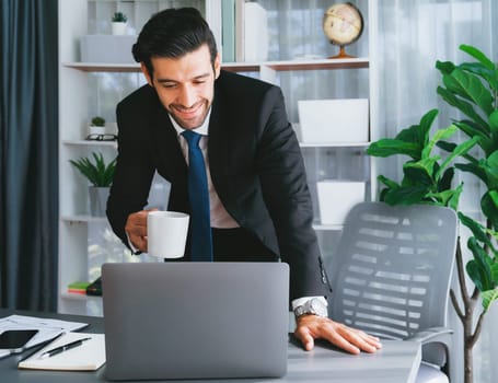 Modern professional businessman at modern office desk using laptop to work with coffee in his hand. Diligent office worker working on computer notebook in his office work space. fervent