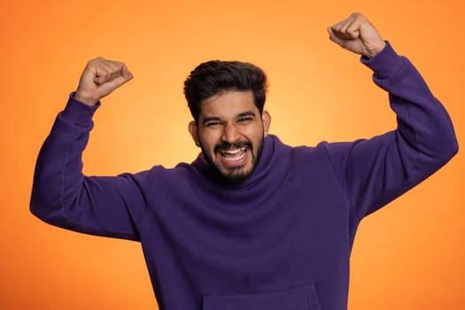 Happy joyful indian man shouting, celebrating success winning, birthday, lottery jackpot goal achievemen, good news, victory, watching TV championship. Young hindu guy on orange studio wall background