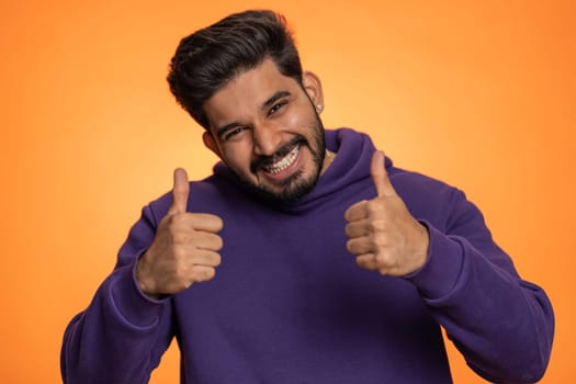 Like. Indian young man in hoodie raises thumbs up agrees with something or gives positive reply recommends advertisement likes good. Handsome bearded hindu guy isolated on orange studio background