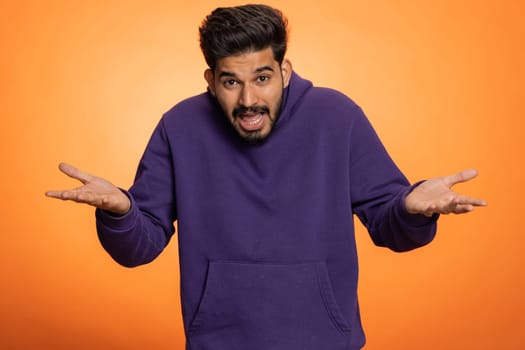 What Why. Sincere irritated indian man raising hands in indignant expression asking reason of failure demonstrating disbelief irritation by trouble. Handsome confused hindu guy on orange background
