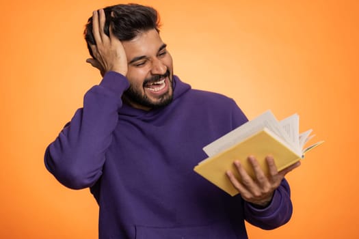 Happy indian man reading funny interesting fairytale story book, leisure hobby, knowledge wisdom, education, learning, study, wow. Handsome hindu guy isolated alone on orange studio wall background