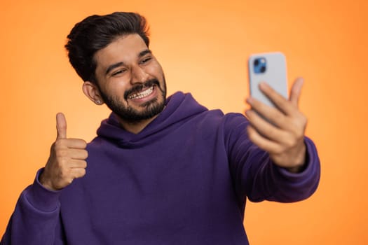 Indian man traveler blogger, taking selfie on smartphone, communicating video call online with family or friends, live translation stream. Handsome hindu guy isolated on orange background, indoors