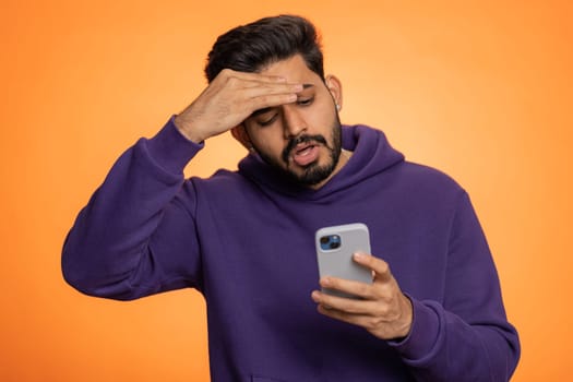 Indian man use mobile cell phone typing browsing, loses becoming surprised sudden lottery results, bad fortune, loss, fail. Handsome hindu guy isolated alone on orange studio wall background, indoors