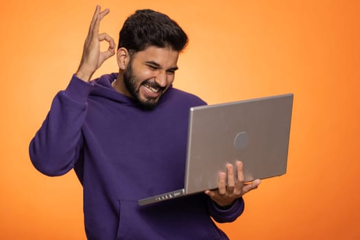 Excited happ young indian man typing on laptop computer, working on project, enjoying results, winning lottery, celebrating success online shopping, win, okay. Hindu guy isolated on orange background