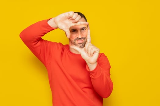 Bearded Hispanic man in his 40s wearing a red sweater making a frame with his fingers, isolated on yellow background. Creativity concept