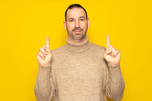 Bearded Hispanic man in his 40s wearing a beige sweater in a friendly attitude pointing up with his index fingers, isolated on yellow studio background