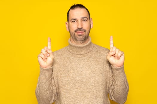 Bearded Hispanic man in his 40s wearing a beige sweater in a friendly attitude pointing up with his index fingers, isolated on yellow studio background