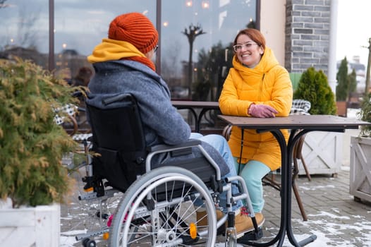 Two girlfriends in a cafe on a street terrace in winter. Woman in a wheelchair