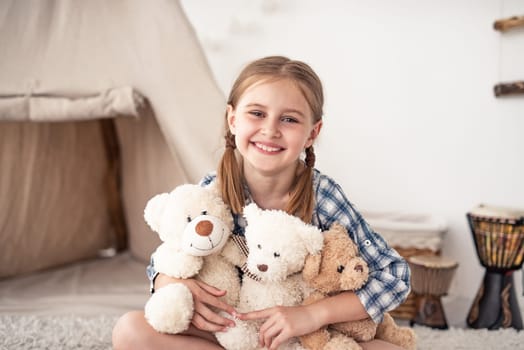 Little girl hugging plush teddies and smiling at camera sitting on floor in playroom