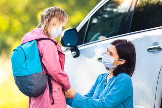 Schoolgirl with mother in protective masks at car parking before classes during coronavirus pandemic