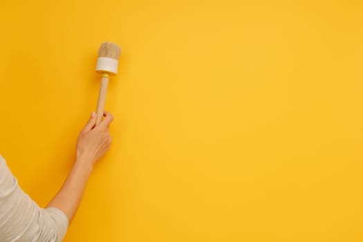 Woman hand holding wooden paint brush on yellow background with copy space. Renovation and improvement equipment. Working tool for house design project and improvement
