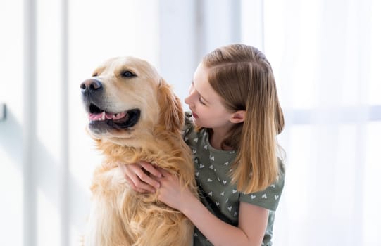 Beautiful child girl hugging golden retriever dog and looking at him indoors. Kid petting purebred doggy pet at home