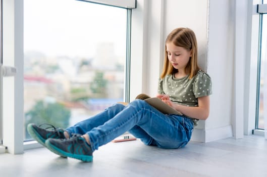 Preteen girl sitting on windowsill and reading paper book with daylight. Beautiful kid schoolgirl learning doing homework at home