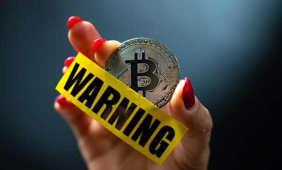 Hand holding bitcoin virtual currency coin and yellow caution tape with word warning. Concept of economy and money instability