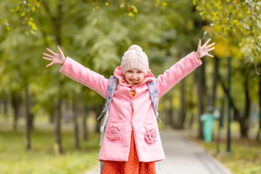 School girl with backpack looking at camera and smiling with hands up at autumn park. Happy preteen child having fun oudoors