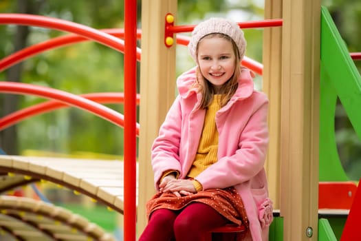 Pretty girl kid sitting on playground at autumn day outdoors, looking at camera and smiling. Female child happy at park