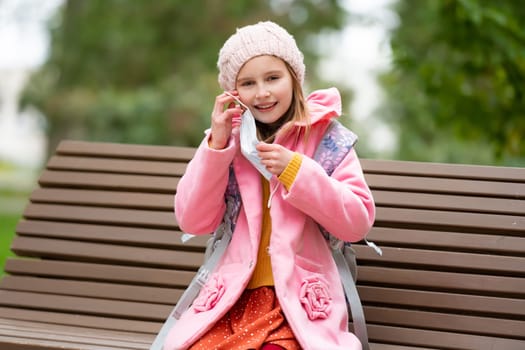 School girl kid wearing medicine mask at autumn park outdoors after lessons. Pretty female child cares about safety at pandemic coronavirus time