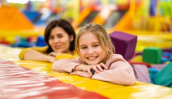 Pretty girls family together on colorful trampoline at playground park smiling. Beautiful kid daughter and her mother happy during active entertaiments indoor