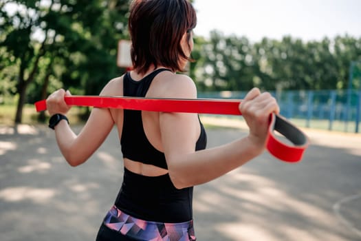 Young girl woman exercising outdoors with rubber elastic band doing training for her back. Closeup view of active fitness with additional sport equipment outside