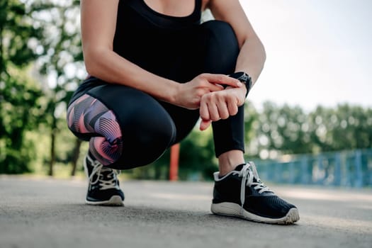 Young girl woman wearing black sportwear sitting at stadium and looking at fitness tracker on her hand during morning workout. Female person control sport progress with smart watch
