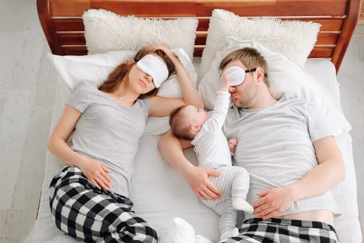 Family with child lying in the bed together in eye masks and sleeping. Young mother and father hugging toddler kid and resting in the bedroom
