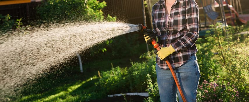 Caucasian woman gardener in work clothes watering the beds in her vegetable garden on sunny warm summer day. Concept of working in the garden