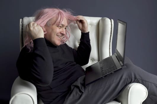 A man in a pink wig sits in a white chair and communicates via video link on a laptop. Side view