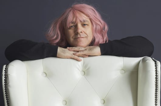 Portrait of a man in a pink wig, who is leaning on a white chair, looking at the camera, posing. close-up.