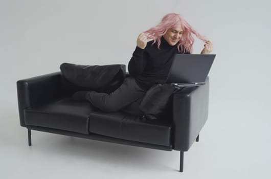 Portrait of a smiling man in a pink wig, sits on a sofa, communicates via video link on a laptop. White background.