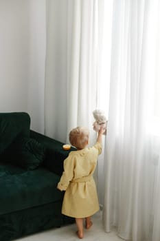 A little boy in a yellow coat plays with a soft toy in the living room.