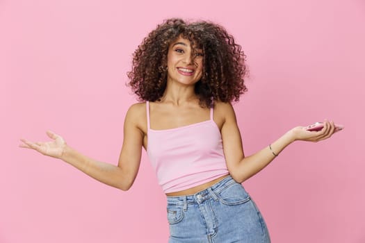 Woman blogger holding phone with curly hair in pink top and jeans poses on pink background, copy space, technology and social media. High quality photo