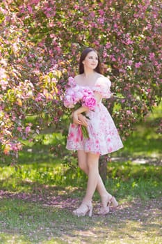 Pretty girl, stands with a large bouquet of peonies, near pink blooming garden, on a sunny day, looking away Copy space. Vertical