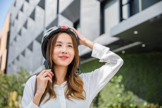 Close up smiling woman wearing helmet exercise outdoors with bicycle on street, Asian young businesswoman putting biking helmet for safety prepared cyclists around building go to work, Bike commuting